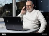 grey-haired-male-talking-on-mobile-phone-whilst-working-at-a-laptop-in-a-home-office-2H7X22P