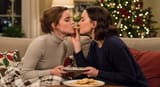 American_sitcom_lighting_of_two_women_Emma_Watson_disgusted_expression_light_brown_hair_wearing_a_form_fitting_turtle_neck_sitting_on_a_couch_with_Daisy_Ridley_dark_brown_hair_both_passionately_kissing_a_cookie_Emma