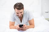 53983064-happy-man-using-mobile-phone-while-lying-on-bed-at-home
