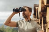 mid-adult-man-looking-through-binoculars-against-sky-in-forest-BOYF01564