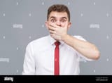 portrait-of-young-shocked-man-in-white-shirt-and-tie-standing-closing-his-mouth-with-hand-and-looking-at-camera-with-shocked-face-and-big-eyes-indoor-isolated-on-gray-background-2GP1XTH