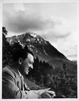 Hitler is talking to someone while overlooking the view in the countryside in Germany. Possibly in the Berchtesgaden area of the Berghof where he liked to go for a rest from Berlin