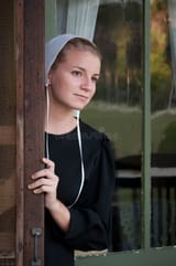 amish-woman-looks-out-screen-door-young-front-porch-green-painted-wood-reflective-glass-background-137435210