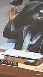 A person with dark skin and dark hair, wearing a dark jacket and a light-colored scarf, is seated at a desk with a &#039;UNITED STATES&#039; nameplate. The person gets up from their chair with a sudden, energetic movement, then _thumb.jpg