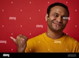 skeptical-indian-man-in-yellow-t-shirt-pointing-with-thumb-and-looking-away-on-red-backdrop-2RTCFN6