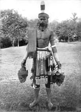 COLLECTIE_TROPENMUSEUM_Portret_van_een_Dajak_krijger_op_Borneo_met_twee_van_hoofddeksels_voorziene_schedels_in_zijn_handen_en_een_kleed_over_zijn_schouder_TMnr_60043379-175754681