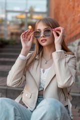 cool-pretty-young-zoomer-woman-with-leather-fashion-white-jacket-jeans-wears-stylish-sunglasses-sits-steps-rest-near-vintage-brick-mall-building_338491-18045