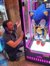 Ian Flynn melodramatically pounds on a glass case containing an oversized Sonic the Hedgehog plush doll